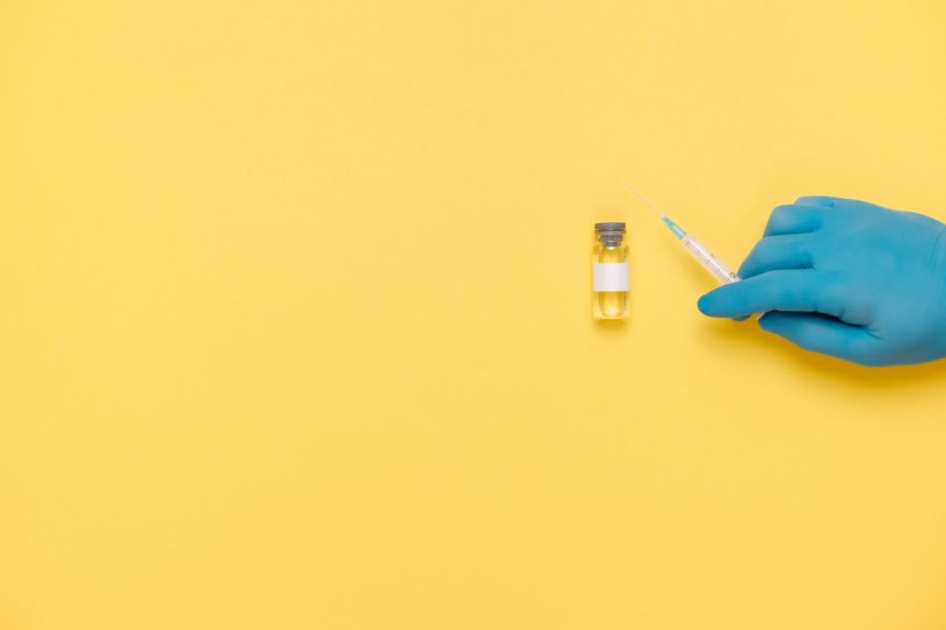 Mão enluvada segura uma seringa próxima a um frasco de vacina sobre fundo amarelo.