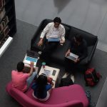 Grupo diverso de adultos colabora usando laptops em uma área de convivência de biblioteca.