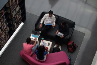 Grupo diverso de adultos colabora usando laptops em uma área de convivência de biblioteca.